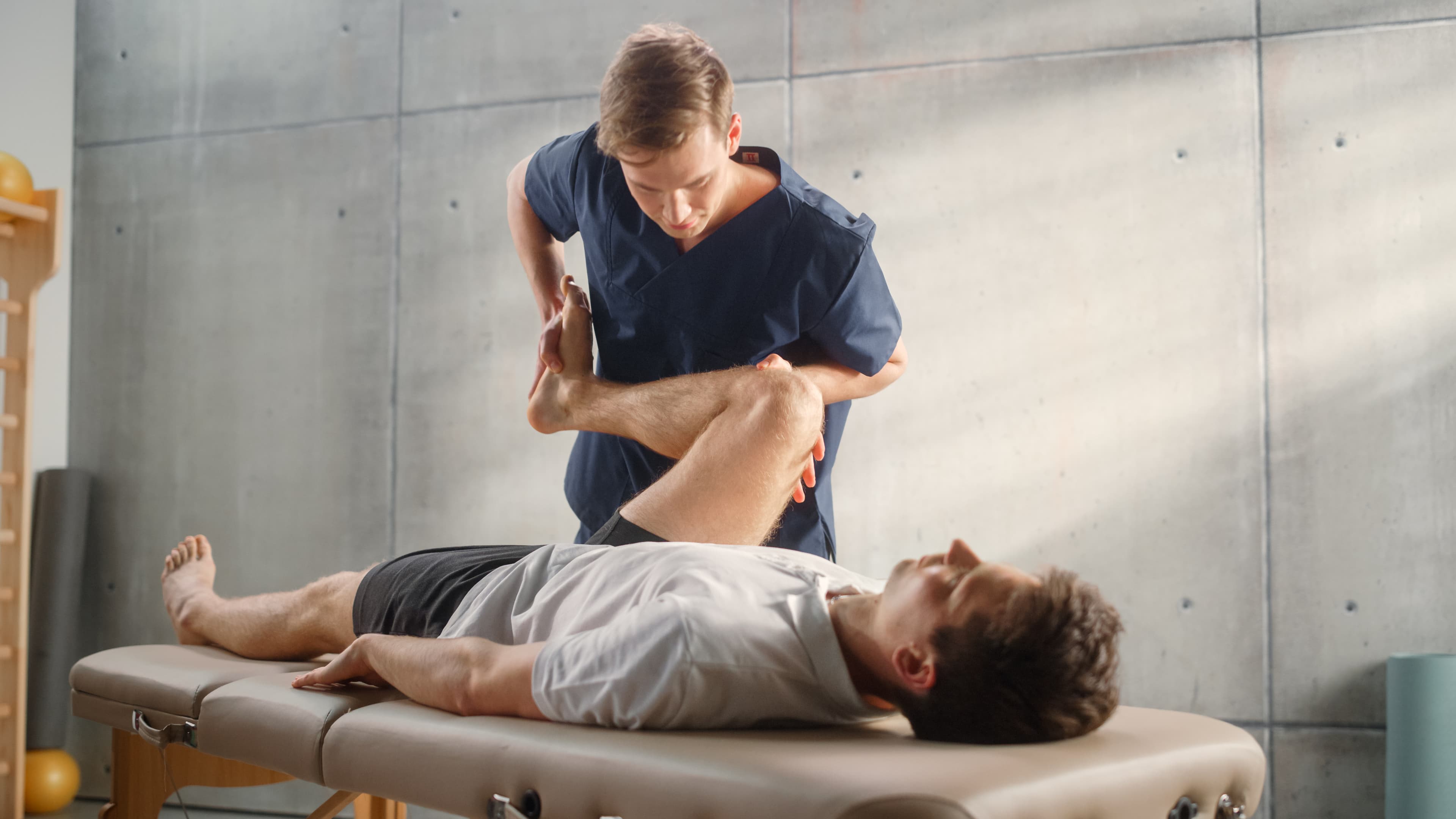 Physical therapist in blue scrubs stretches a patient's leg on a treatment table.