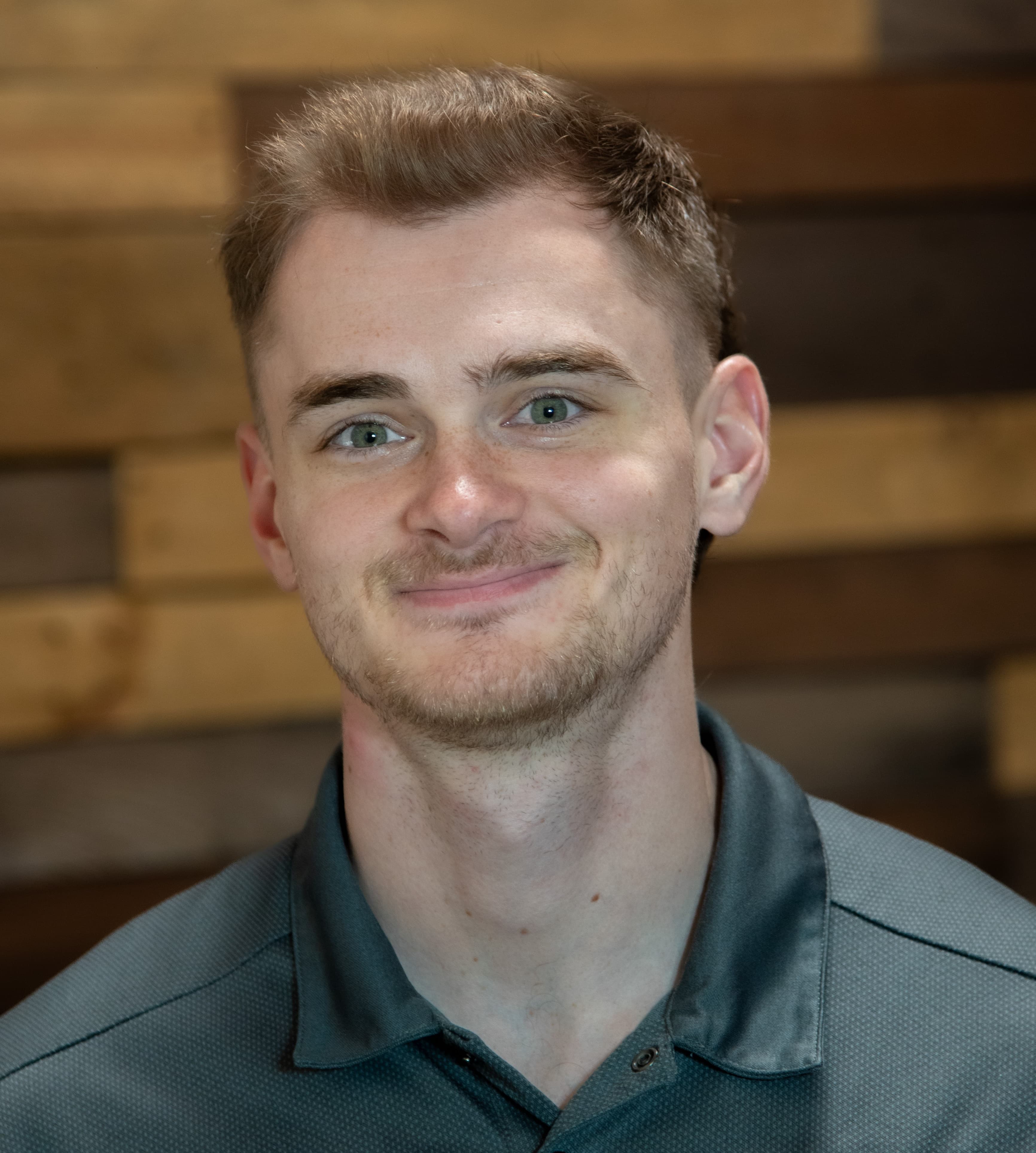 Smiling young man with green eyes and light stubble wearing a grey polo shirt indoors.